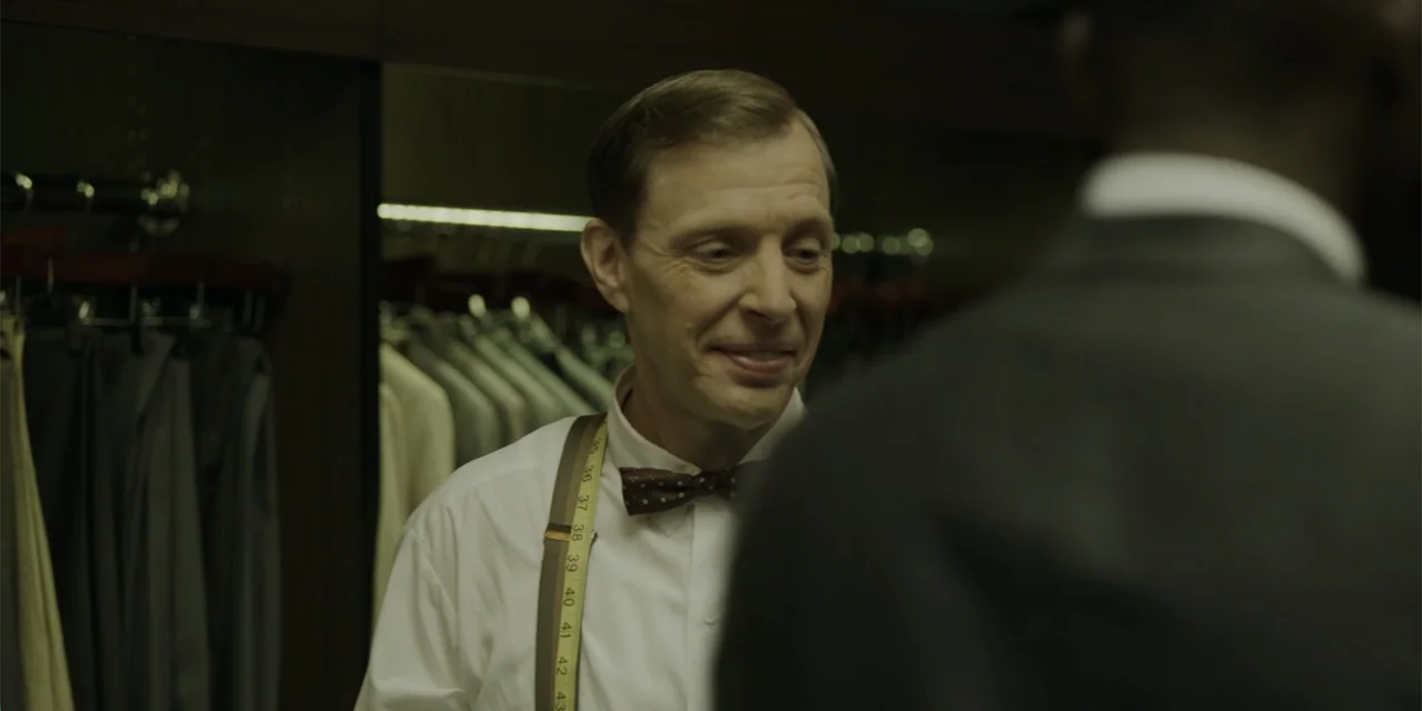 Green Book (2018) movie still: medium — A tailor shop filled with racks of suits and dark wooden wall paneling. A middle-aged man in a dress shirt…