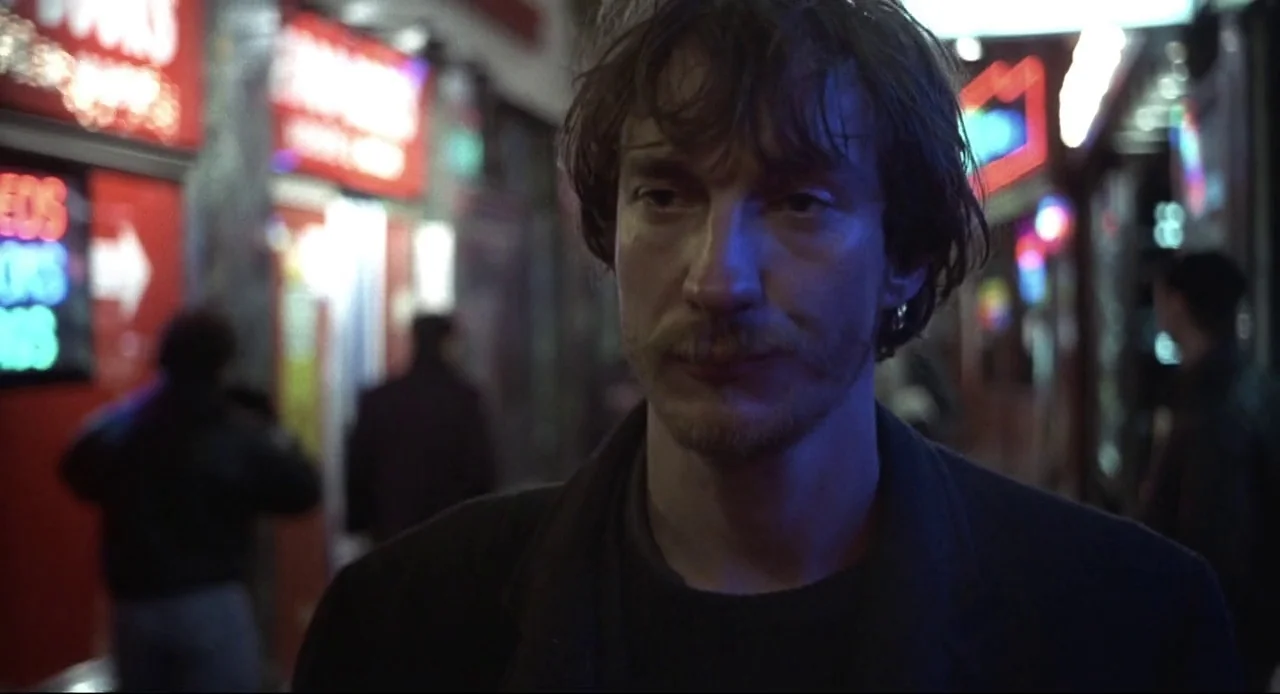 Naked (1993) movie still: closeup — A nighttime urban scene with red and blue neon signs reflecting on the surroundings. A man with messy hair and…