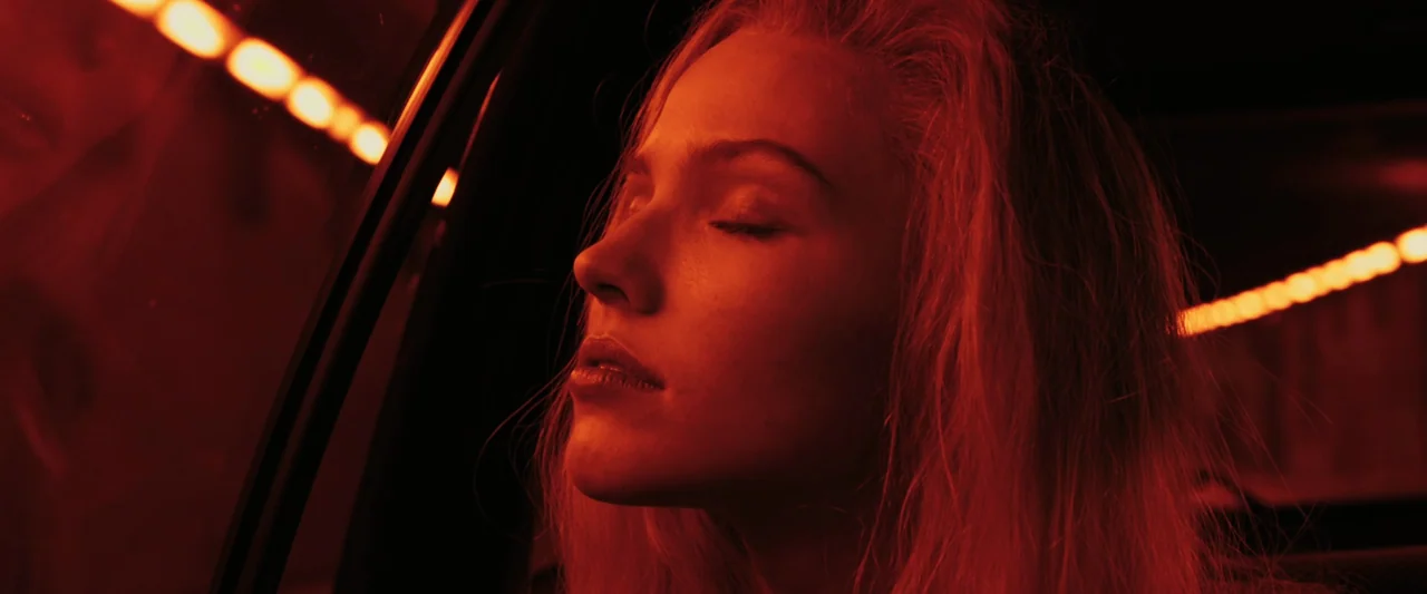 Anna (2019) movie still: closeup — A woman sits in the back of a moving vehicle at night. The interior is dark while amber light blurs against the…