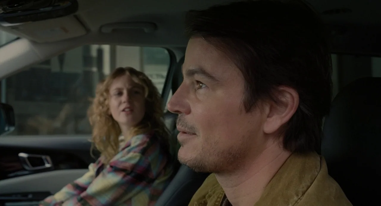 Trap (2024) movie still: medium — A man and woman are seated inside a car during the day. The man drives while the woman in the backseat looks…