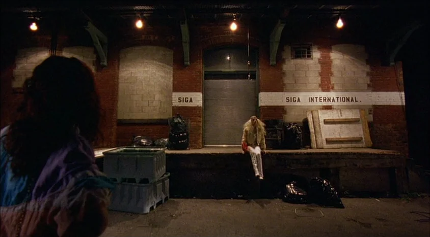 Hedwig and the Angry Inch (2001) movie still: wide — A dark, brick-walled industrial loading dock features a large metal roll-up door. A person in a…