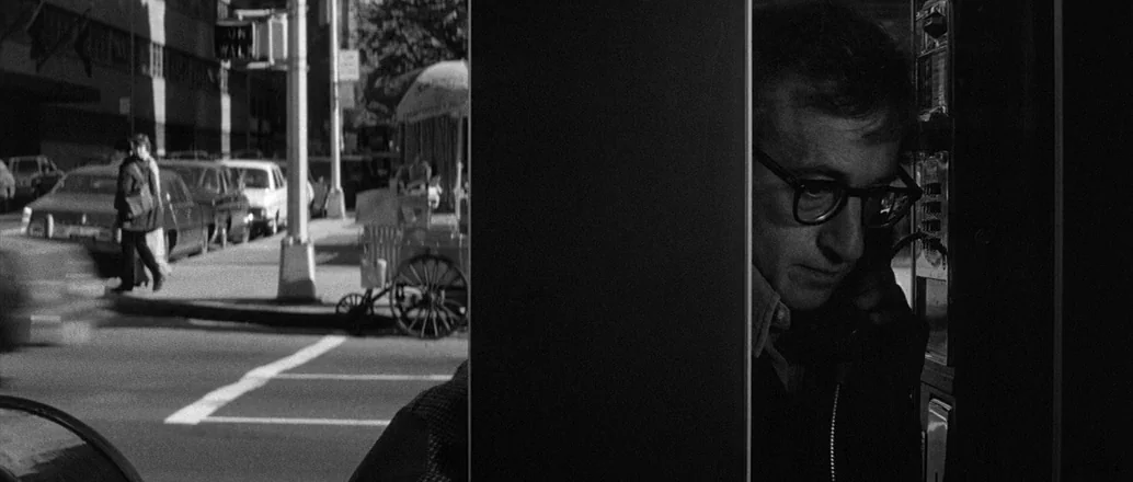 Manhattan (1979) movie still: medium — A man stands inside a narrow metal phone booth on a busy city sidewalk. Pedestrians walk past parked cars on…