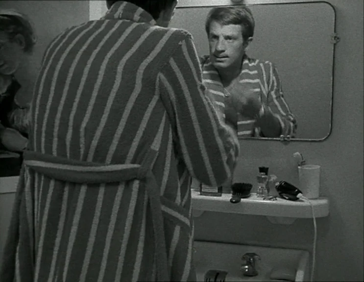 À Bout De Souffle (Breathless) (1960) movie still: medium — A man stands before a wall-mounted mirror in a plain tiled bathroom. He wears a striped…