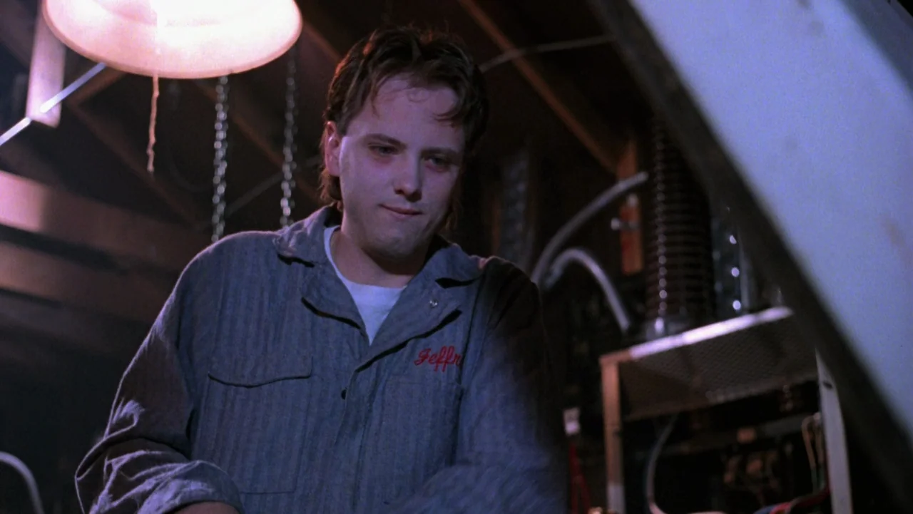 Frankenhooker (1990) movie still: medium — A dimly lit workshop interior contains metal structures and hanging chains in the background. A young man…