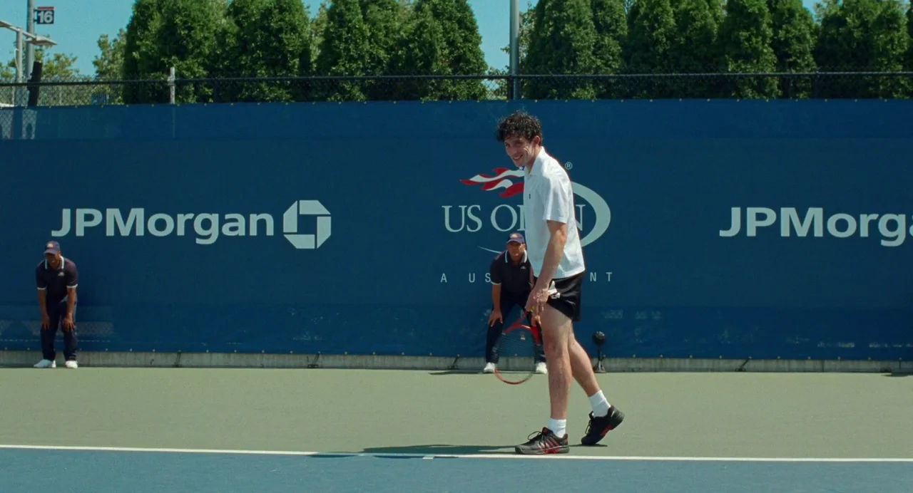 Challengers (2024) movie still: fullbody — An outdoor hard-surface tennis court features blue backdrops with corporate branding logos. A male tennis…