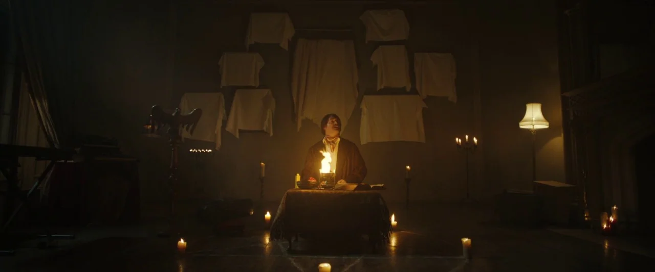 Extra Ordinary (2019) movie still: wide — A dimly lit room features white cloths hanging on a back wall. A man sits at a central table with a small…