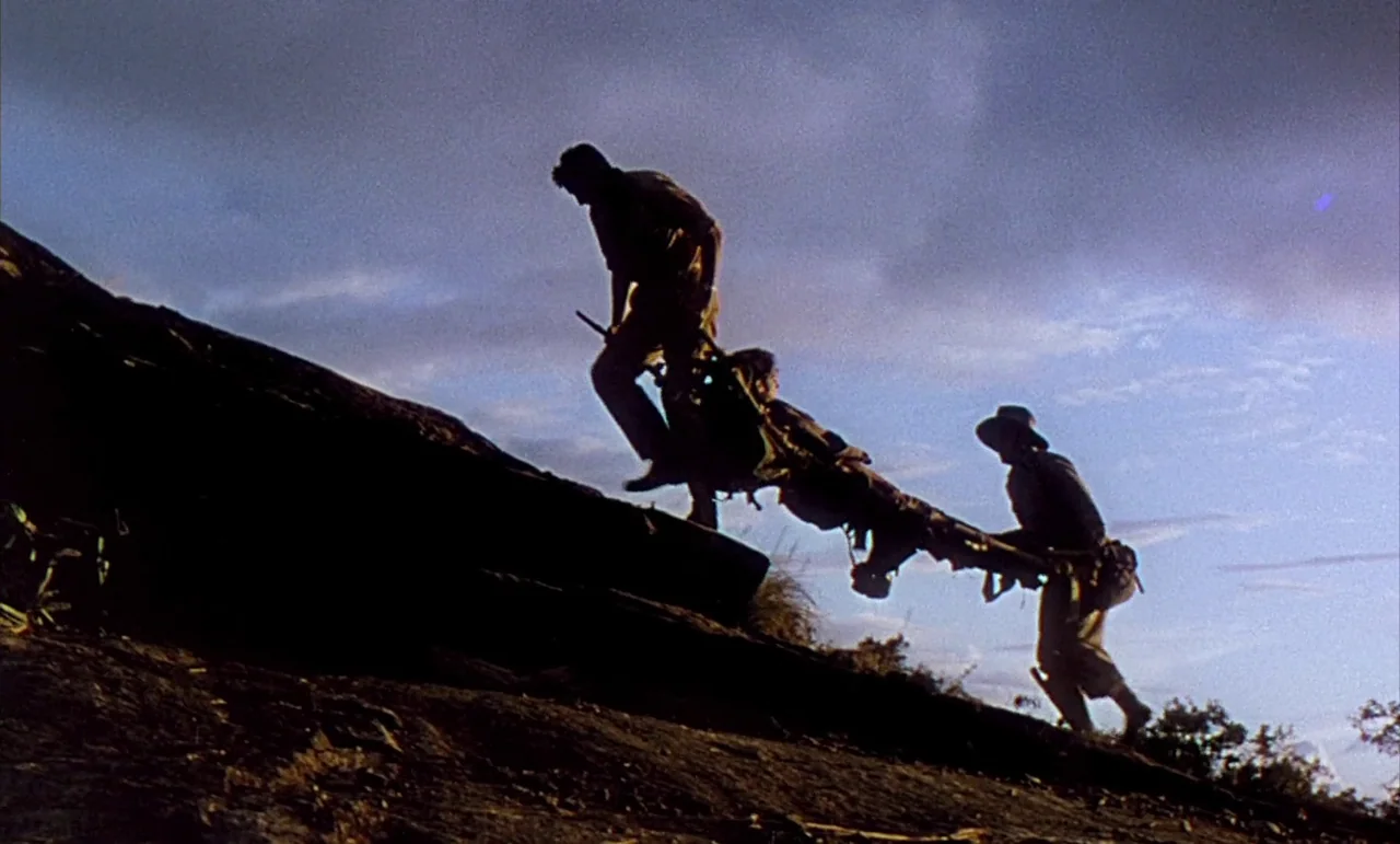 The Purple Plain (1954) movie still: wide — A steep rocky incline under a cloudy sky during the day. Two men in uniform carry a wounded person on a…