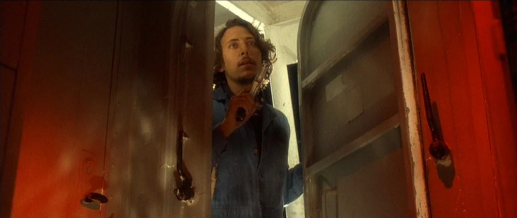 The Usual Suspects movie still: medium — A cramped and dark train car interior with industrial metal doors framed in red light. A young man stares…