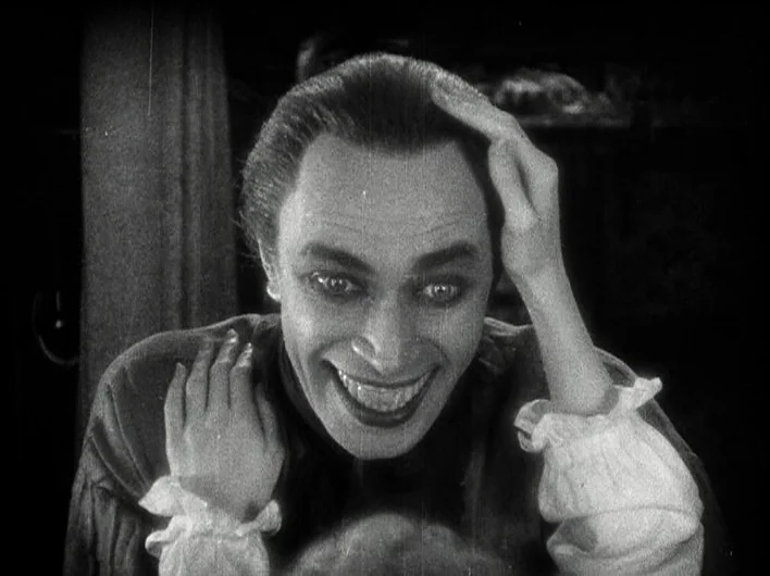 The Man Who Laughs (1928) movie still: closeup — An interior space is dark with only the man's face clearly visible. The subject pulls his hair back…