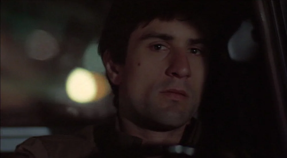 Taxi Driver (1976) movie still: closeup — The interior of a moving vehicle at night with a blurred urban backdrop. A young man with dark hair…