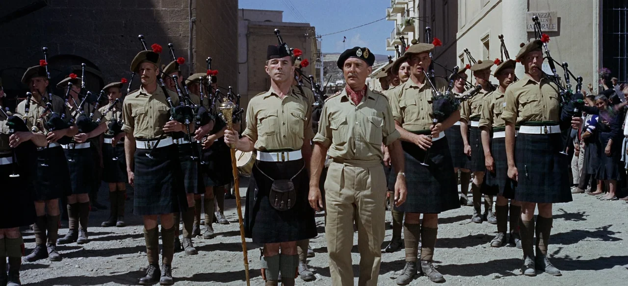 Patton (1970) movie still: fullbody — A group of Scottish bagpipers in military uniform marches down a narrow dusty street. The men carry bagpipes…