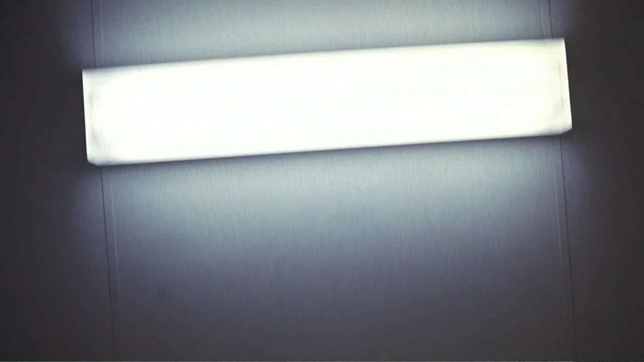 The Forever Prisoner (2021) movie still: establishing — A plain gray wall features a rectangular fluorescent light fixture mounted horizontally. The…