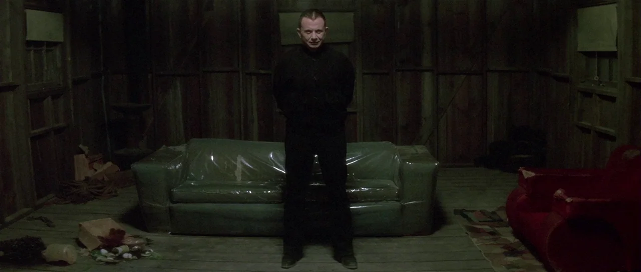 Lost Highway (1997) movie still: fullbody — A dimly lit cabin interior features wood-paneled walls and a sofa wrapped in plastic. A man in dark…