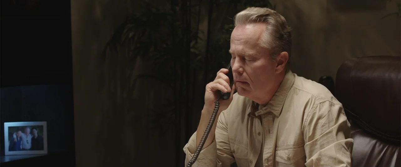 Presagios (2024) movie still: medium — A dimly lit office features dark walls and a potted plant in the background. A man sits at a desk while…