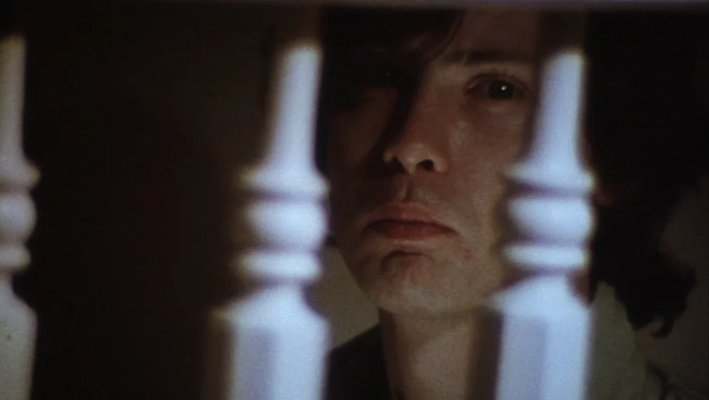 Martin (1977) movie still: closeup — A man peers through the vertical wooden balusters of an indoor staircase at night. The foreground is partially…
