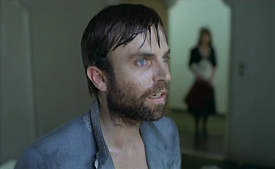 Possession (1981) movie still: closeup — A man stands in the foreground of a dimly lit, nondescript domestic hallway. A woman remains out of focus…