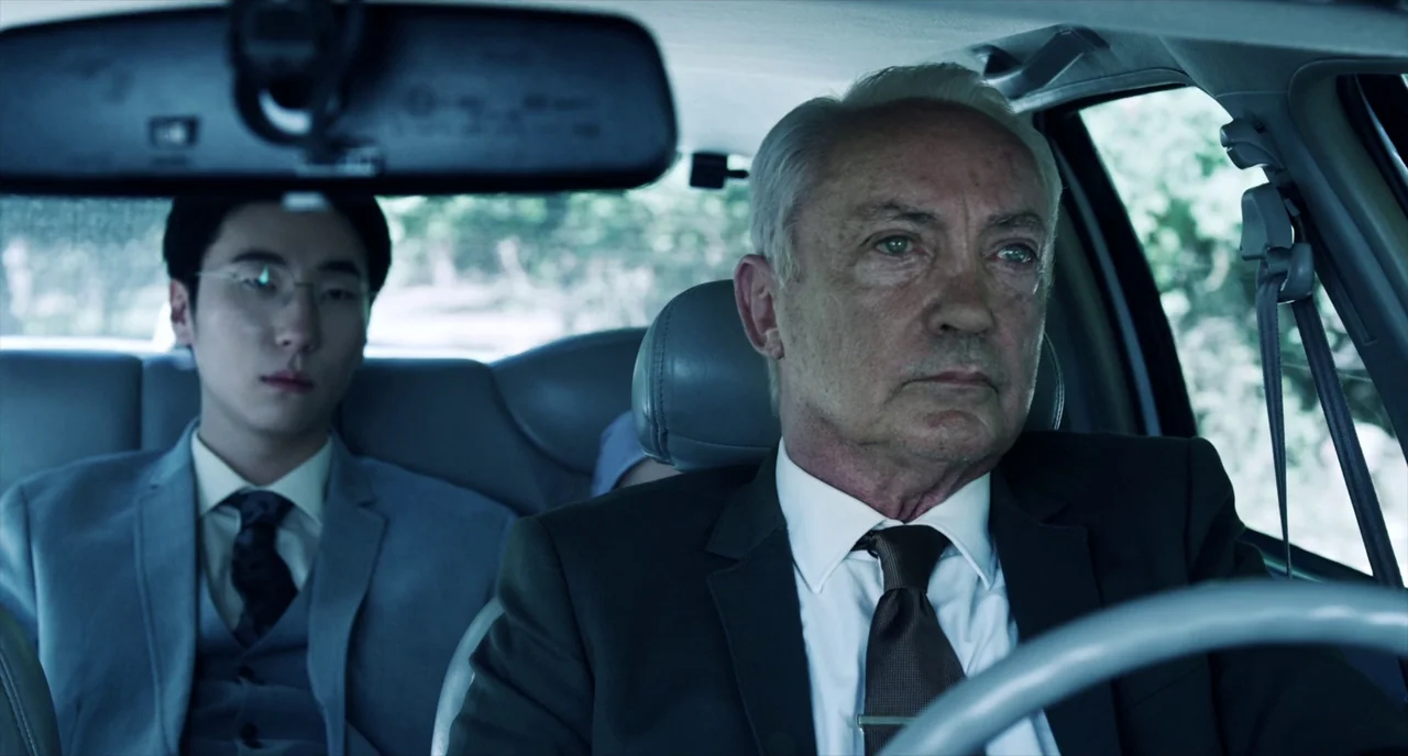 Brawl in Cell Block 99 (2017) movie still: medium — The interior of a car features two men in formal business suits. An elderly driver watches the…