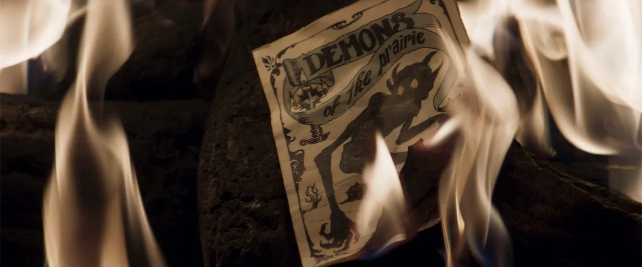 The Wind (2005) movie still: closeup — A piece of aged paper with printed devilish artwork sits atop charred wooden logs. The edges of the paper…