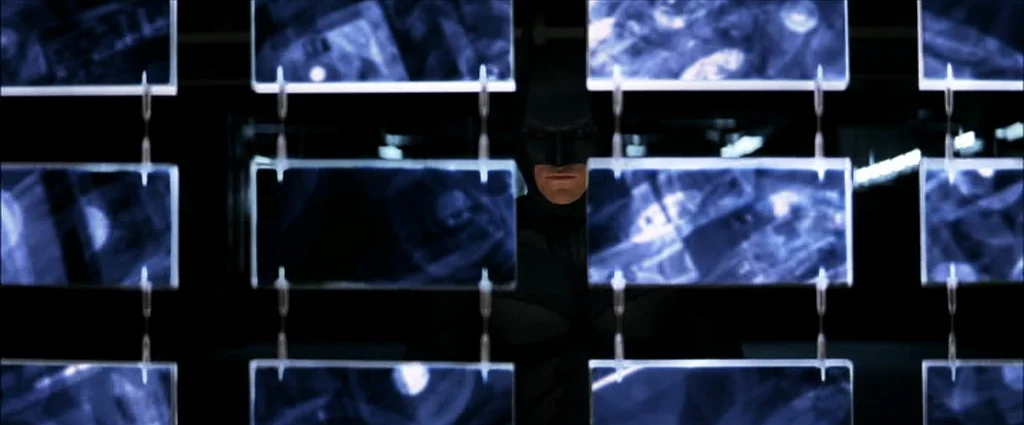 The Dark Knight (2008) movie still: medium — A dark, high-tech interior space featuring a wall of suspended translucent blue monitors. Batman stands…