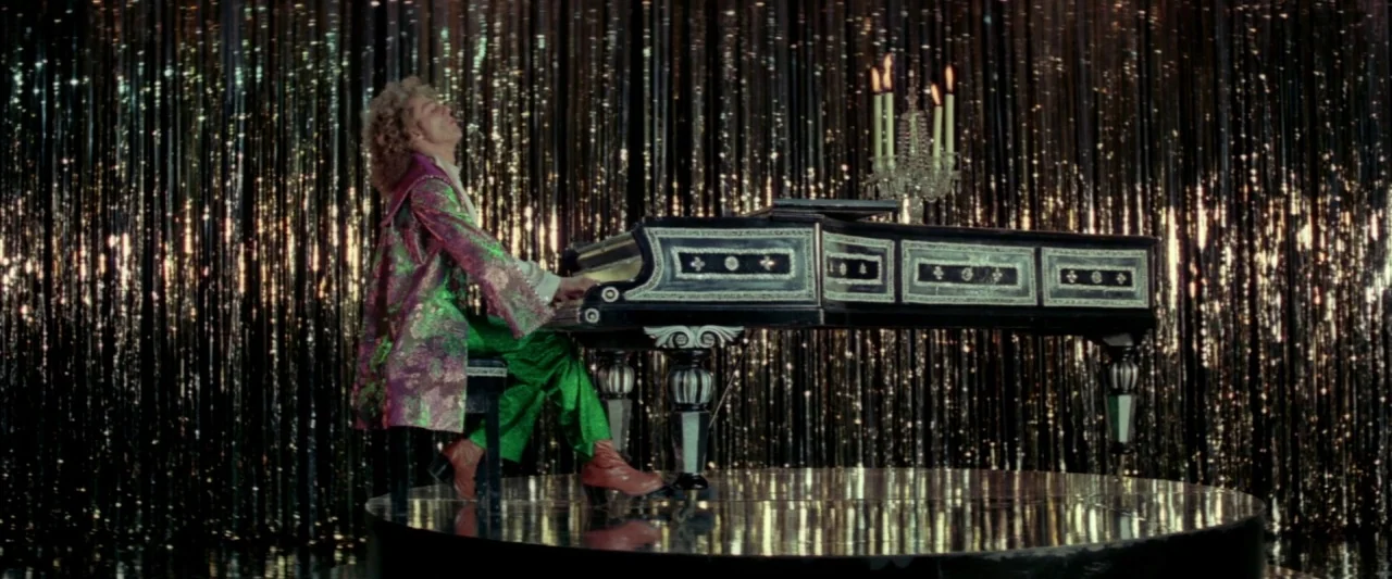 Lisztomania (1975) movie still: fullbody — A circular stage with a black grand piano sits before a wall of metallic fringe. A man in a patterned…