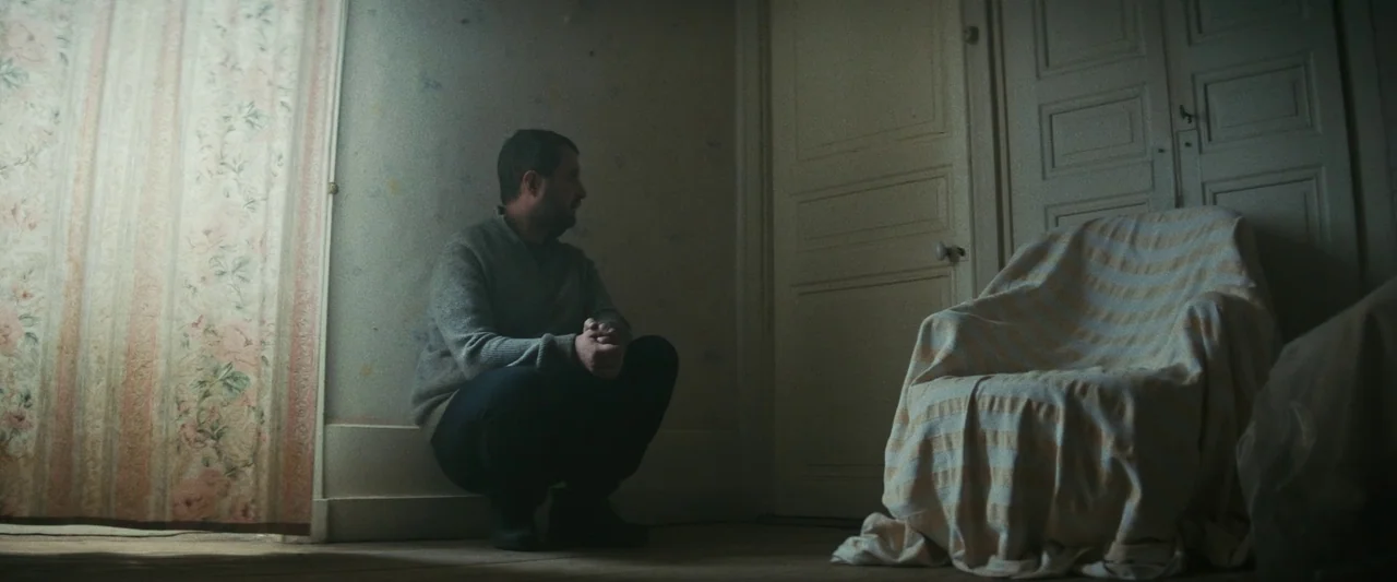 Vincent Must Die (2023) movie still: medium — A quiet, sparse room features a patterned curtain, peeling wallpaper, and a fabric-covered chair. A…