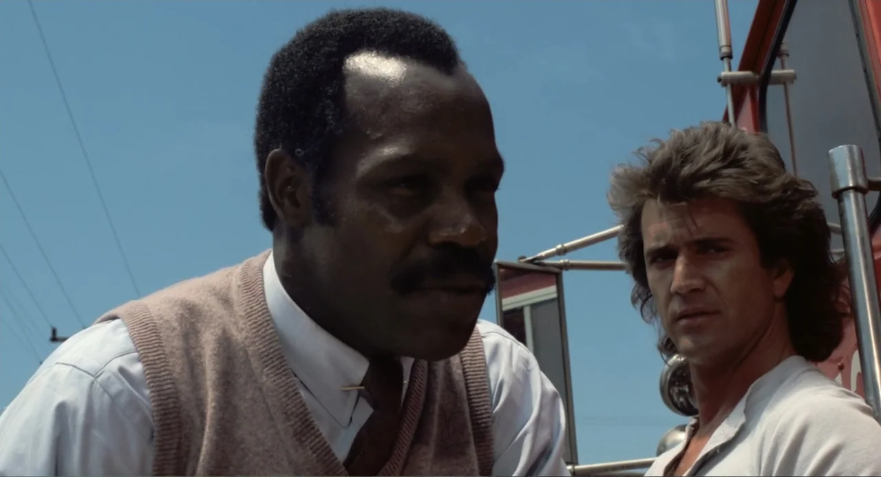 Lethal Weapon (1987) movie still: medium — An exterior view from within a vehicle cabin against a bright blue sky. Two men sit side by side while…