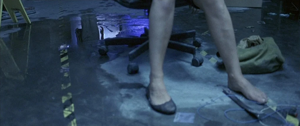 Boarding Gate (2007) movie still: closeup — A dark industrial concrete floor is covered in shallow pools of standing water. A rolling office chair…