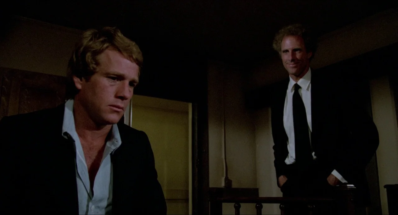 The Driver (1978) movie still: medium — A dimly lit interior hallway with dark walls and a simple wooden stair railing. A young man sits in the…