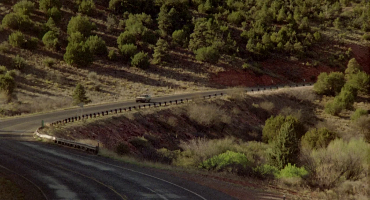 The Karate Kid (1984) movie still: wide — A winding asphalt road cuts through a hilly landscape filled with scrubby vegetation. A lone sedan drives…