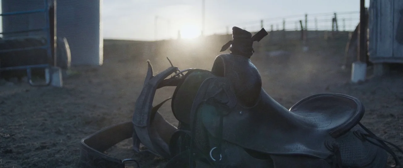 The Rider (2017) movie still: closeup — A leather western saddle sits on the dusty ground of an outdoor livestock pen. A sun-bleached metal grain…