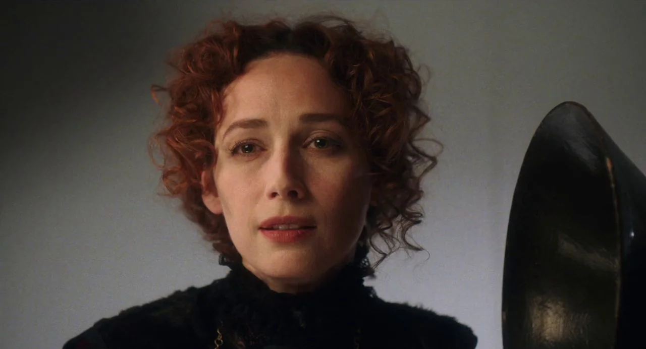 Tesla (2020) movie still: closeup — A woman stands in a neutrally colored room against a plain backdrop. She faces the camera directly with a black…