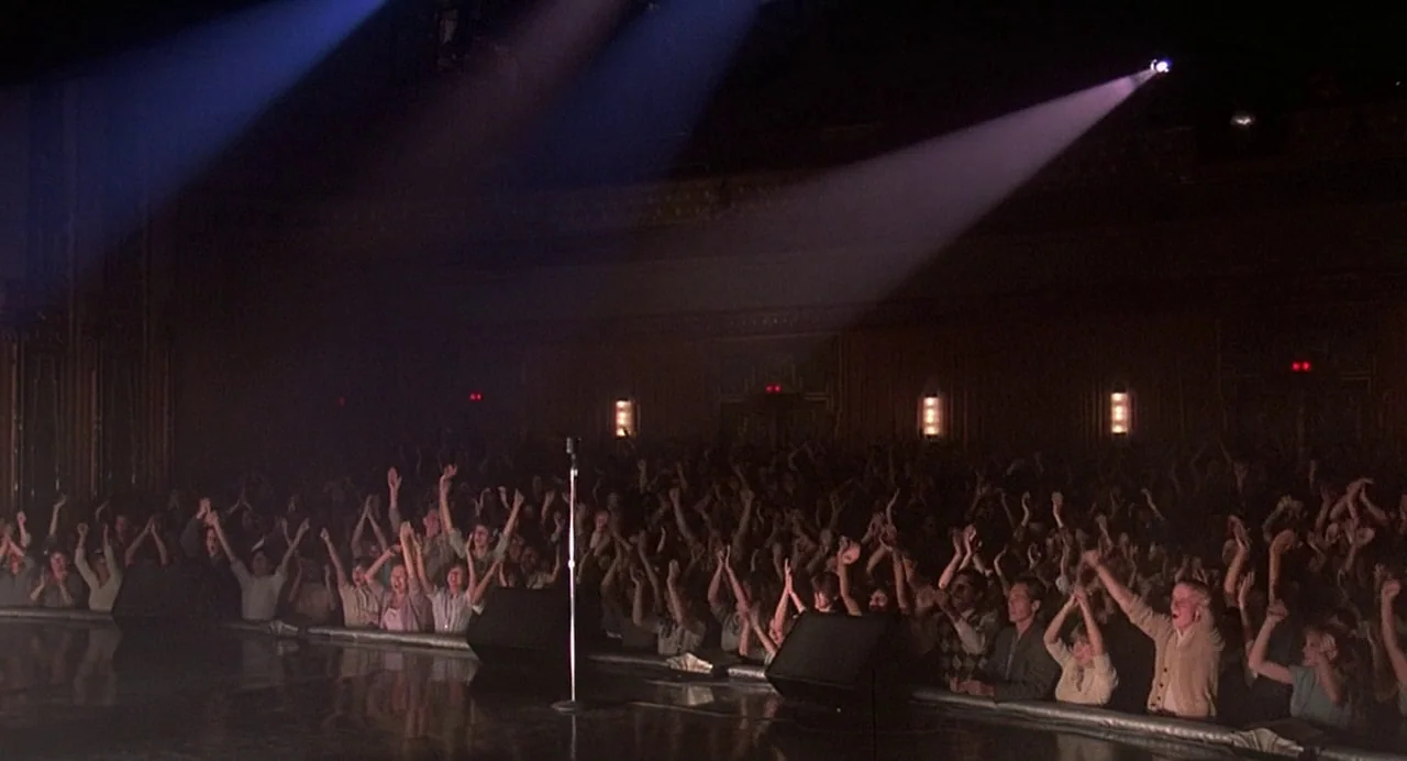 Streets of Fire (1984) movie still: wide — A dark, large auditorium is filled with a seated audience under dramatic stage lighting. Members of the…