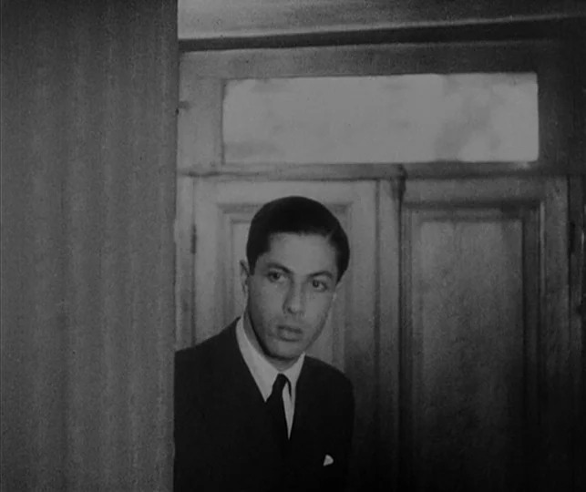 Vampyr (1932) movie still: medium — A man stands partially obscured by a wooden frame in a dimly lit room. He looks toward the side while standing…