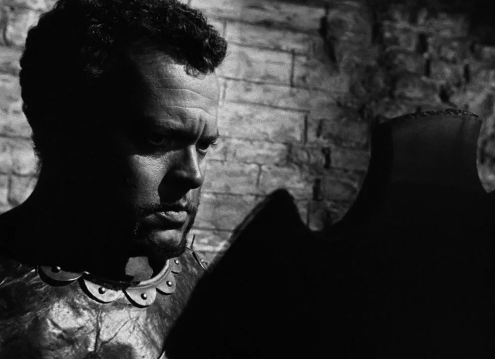 Othello (1952) movie still: closeup — A dark stone wall serves as the backdrop to a man in armor. The man looks toward an obscured object in the…