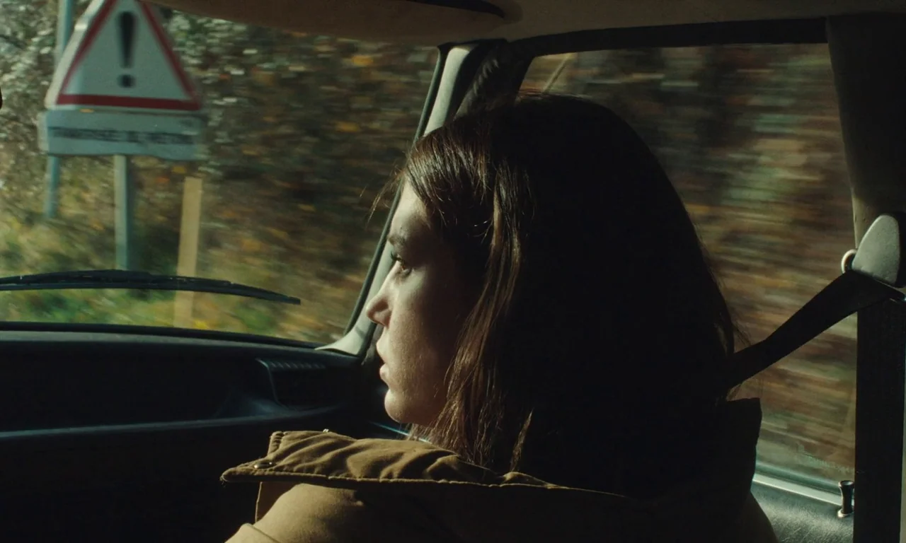 Passages (2023) movie still: medium — A woman sits in the passenger seat of a car moving past a road sign. The car interior is dark and the view…