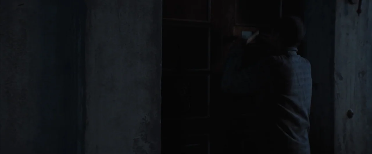 My Neighbor Adolf (2022) movie still: medium — A dark stone wall sits beside a tall, dark wooden door at night. A man stands facing the door while…