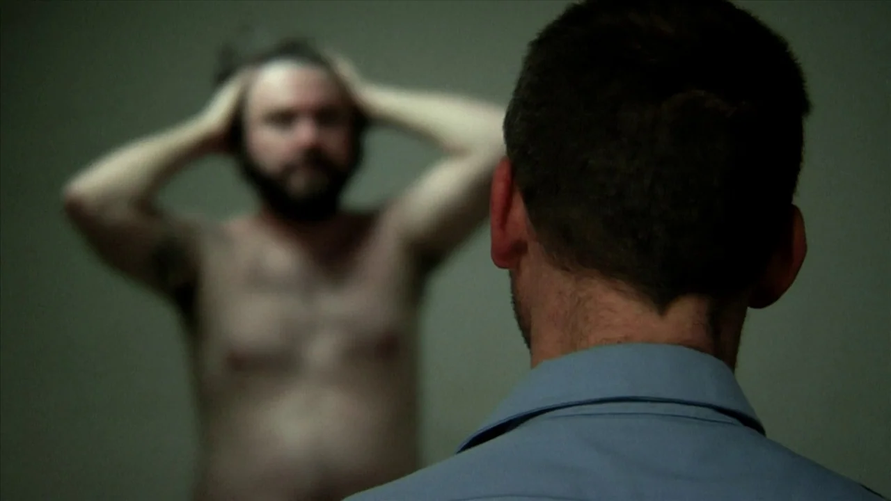 A Horrible Way To Die (2010) movie still: medium — An austere room with plain, featureless walls serves as a space for observation. One man stands…