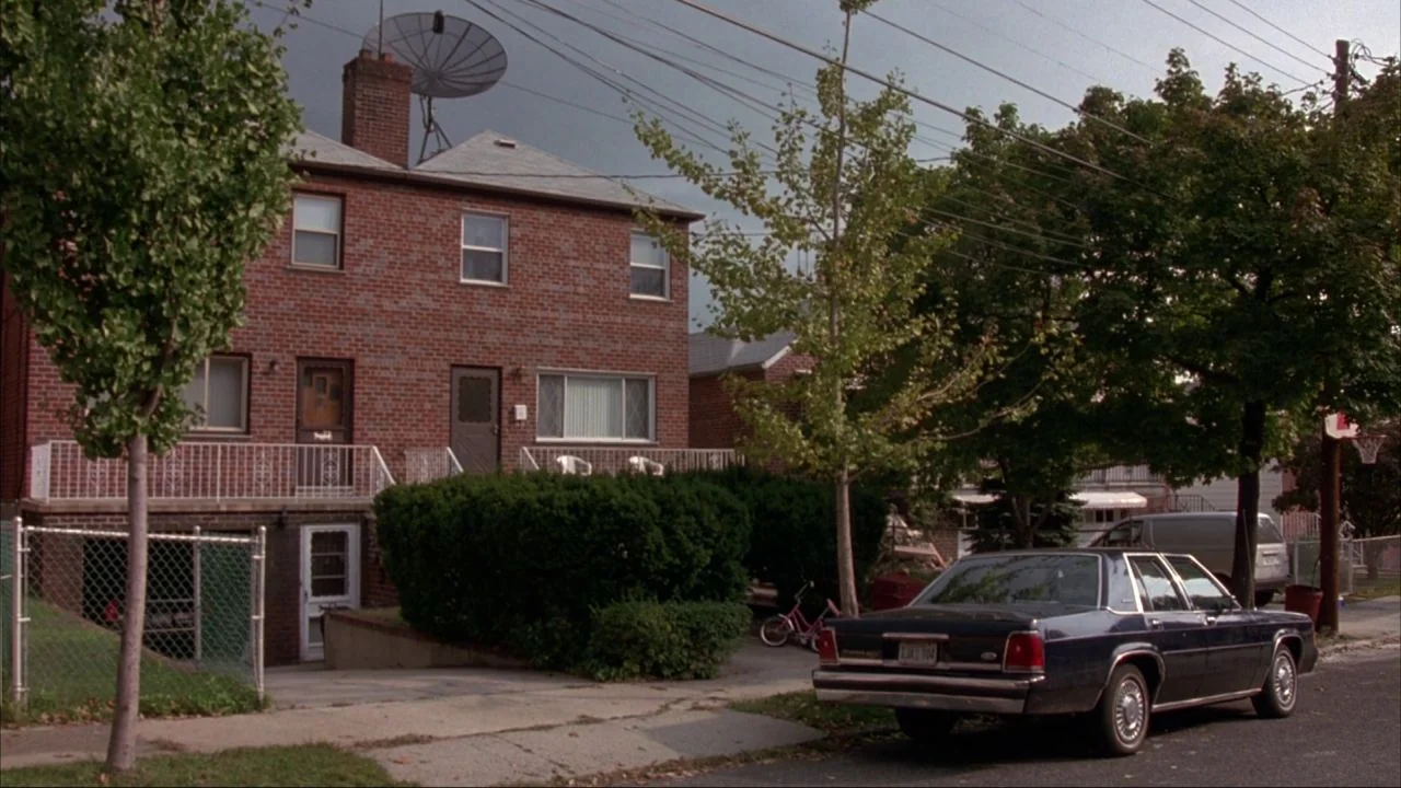 Bad Lieutenant (1992) movie still: wide — A two-story red brick residential building stands behind a thick hedge and sidewalk. A dark sedan is…