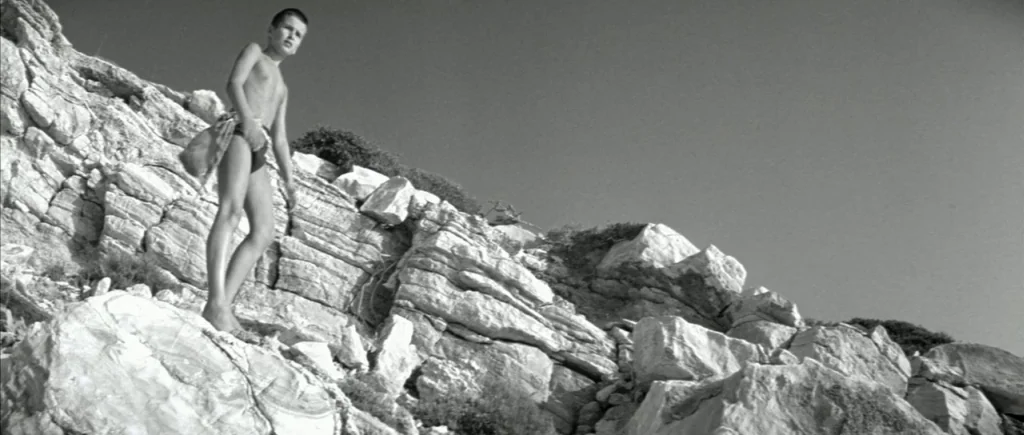 The Big Blue (1988) movie still: fullbody — A steep, sun-drenched coastal hillside composed of jagged, layered white rocks. A young boy stands…