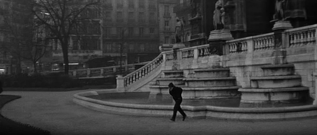 The 400 Blows (1959) movie still: wide — A stone fountain sits in a large, open public plaza surrounded by city buildings. A lone man in dark…