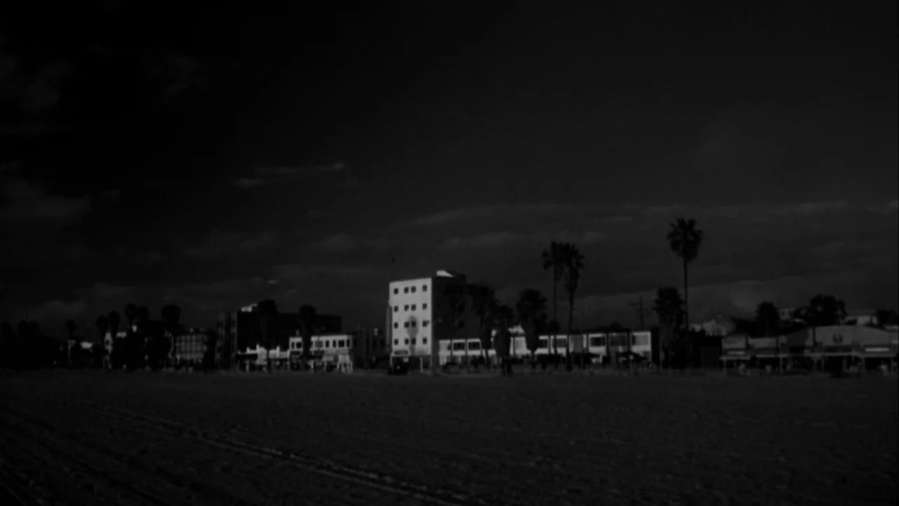 American History X movie still: establishing — A wide view of a coastal beach at night looking toward a line of buildings. Palm trees silhouette…