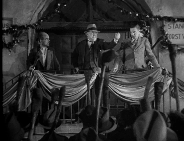 Frankenstein movie still: medium — An exterior balcony decorated with banners and pine garlands overlooks a large gathering. Three men stand on the…