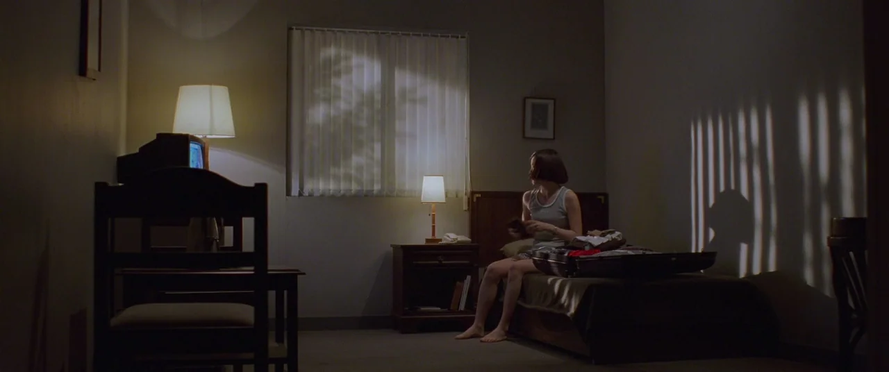 Joint Security Area (2000) movie still: wide — A dimly lit hotel room contains a bed, a suitcase, and two table lamps. A young woman sits on the…