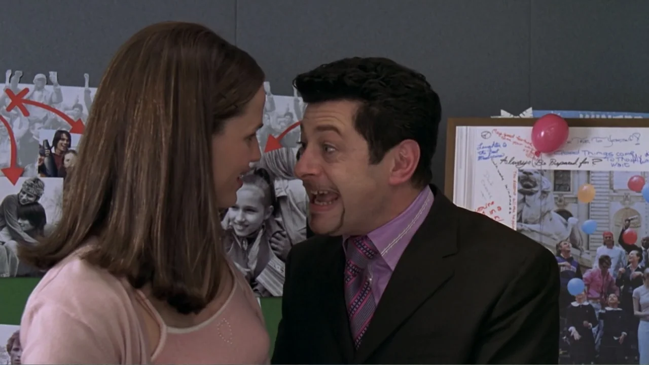 13 Going on 30 (2004) movie still: medium — An interior office space features a dark wall covered with printed photo collages. A man in a suit…