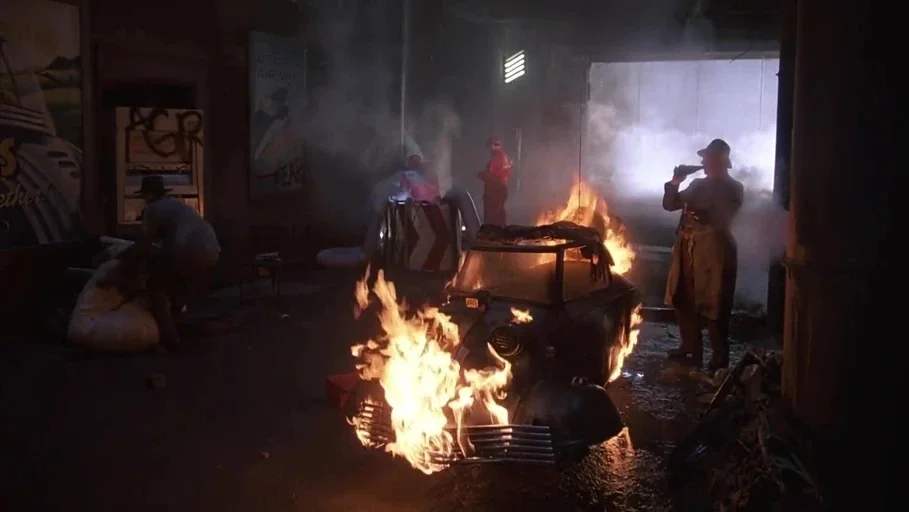 Brazil (1985) movie still: wide — A dimly lit industrial garage filled with thick smoke and several figures present. A vintage automobile sits in…
