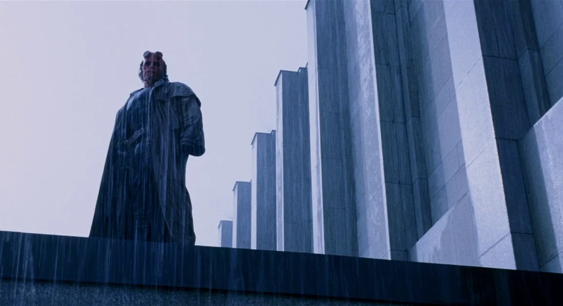Hellboy (2004) movie still: fullbody — A tall stone building facade stands under heavy rain during a dark night. The red-skinned humanoid figure…