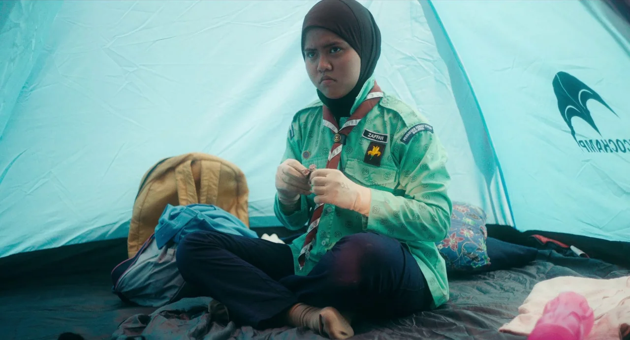 Tiger Stripes (2023) movie still: medium — A person sits inside a light blue nylon camping tent during the day. The subject is wearing a scout…