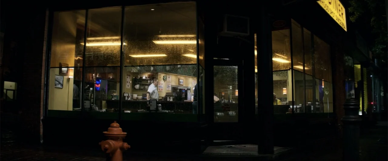 The Equalizer (2014) movie still: wide — A night scene showing a rainy street view of a lit storefront window. A lone employee stands inside the…