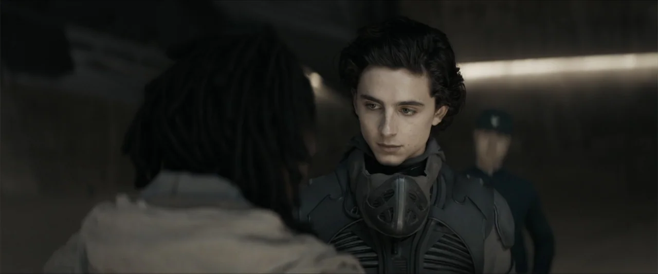 Dune (2021) movie still: medium — A dimly lit industrial space features concrete walls and muted, monochromatic tones. A young man wearing a…