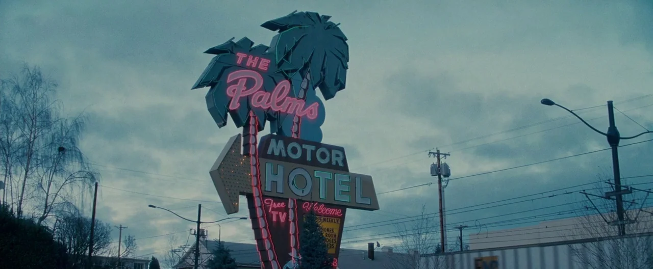 The Burning Plain (2008) movie still: establishing — A retro neon sign stands prominently in front of a gray, overcast sky. Utility poles and bare…